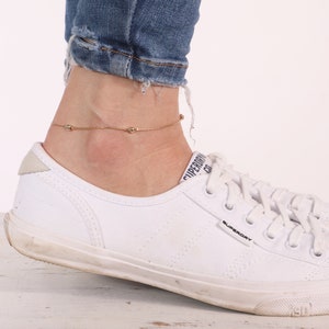 May include: A gold anklet with small round beads on a delicate chain. The anklet is worn on a person's ankle above a white sneaker.