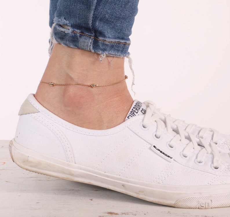 May include: A gold anklet with small round beads on a delicate chain. The anklet is worn on a person's ankle above a white sneaker.