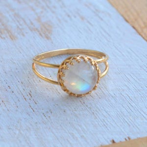 Puede incluir: Un anillo de oro con una piedra de luna redonda e iridiscente engastada en un delicado bisel adornado. El anillo tiene una banda doble.