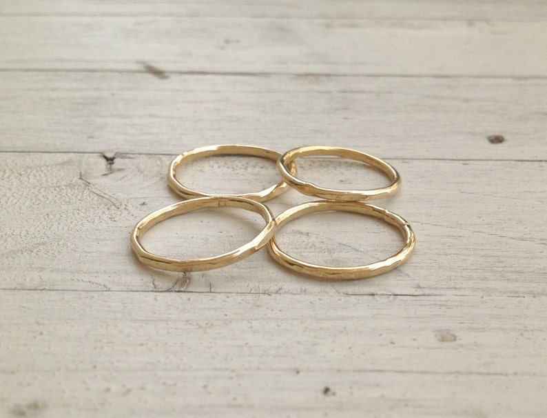 Pu&ograve; includere: Quattro anelli dorati martellati sono disposti su una superficie di legno chiaro. Gli anelli hanno un aspetto strutturato e un design semplice e minimalista. Gli anelli sono circolari e sembrano essere di metallo.