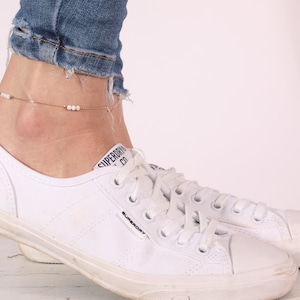 May include: A gold anklet with white beads on a woman's ankle. The anklet is worn with white sneakers and blue jeans.