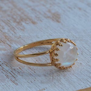 Puede incluir: Un anillo de oro con una piedra de luna blanca engastada en un delicado dise&ntilde;o de corona.