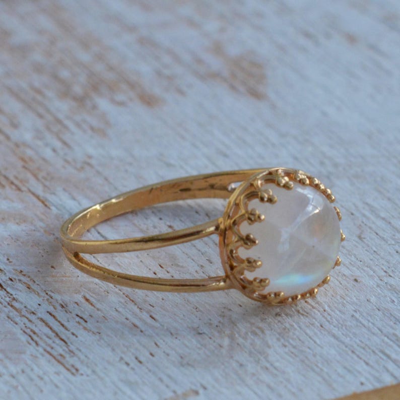 Puede incluir: Un anillo de oro con una piedra de luna blanca engastada en un delicado dise&ntilde;o de corona.