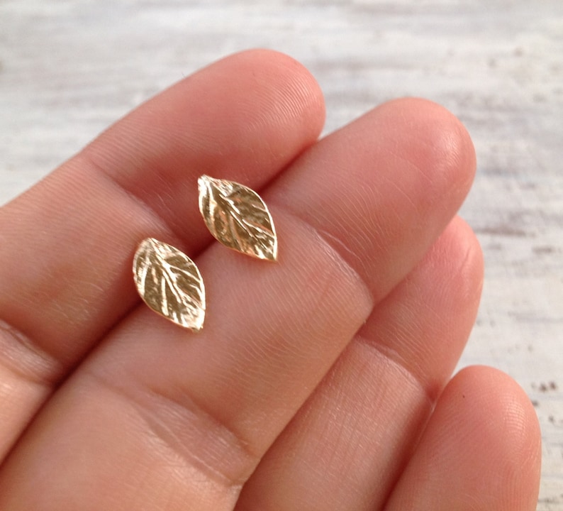 May include: A pair of gold leaf stud earrings. The earrings are small and delicate, and they have a realistic leaf design.
