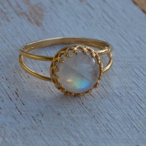 Puede incluir: Un anillo de oro con una piedra de luna redonda e iridiscente engastada en un bisel delicado y adornado. El anillo tiene una banda doble.