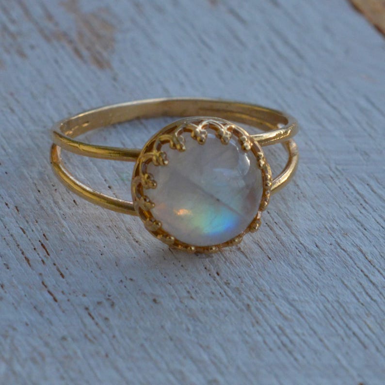 Puede incluir: Un anillo de oro con una piedra de luna redonda e iridiscente engastada en un bisel delicado y adornado. El anillo tiene una banda doble.