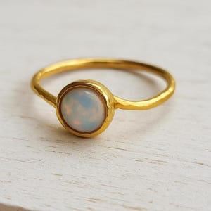 May include: A gold-colored ring with a round, iridescent opal stone. The ring has a simple band and a bezel setting that holds the opal. The opal has a milky white and blue color. The ring is on a white surface.