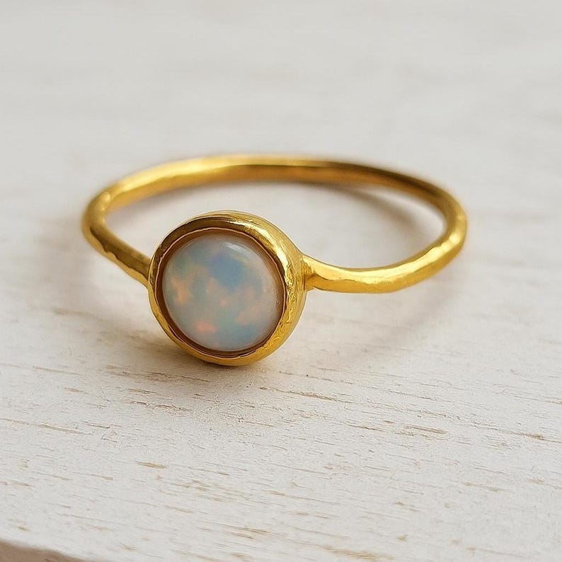 May include: A gold-colored ring with a round, iridescent opal stone. The ring has a simple band and a bezel setting that holds the opal. The opal has a milky white and blue color. The ring is on a white surface.
