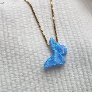 May include: A gold-colored chain necklace with a blue and green opal butterfly pendant. The butterfly pendant has a shimmering, iridescent appearance. The necklace is displayed on a white, textured surface.