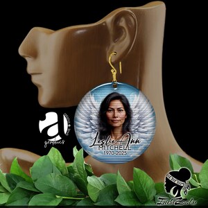 May include: A round, blue and white memorial earring with a photo of a woman with angel wings. The text "Leslie Ann Mitchell 1970-2025" is printed on the earring.