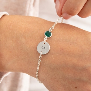 Op de afbeelding: Een zilveren kettingarmband met een ronde bedel met de letter "C" en twee groene edelsteen accenten.
