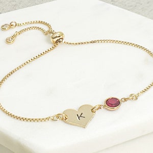 May include: A gold adjustable bracelet with a heart charm engraved with the letter "K" and a red gemstone.