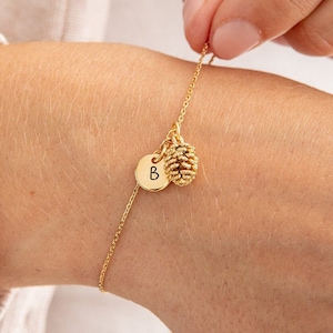 Könnte beinhalten: Ein goldenes Kettenarmband mit einem kleinen Tannenzapfen-Anhänger und einem personalisierten Initialen-Anhänger. Der Initialen-Anhänger ist mit dem Buchstaben "B" graviert.