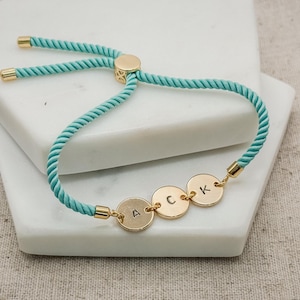 Könnte beinhalten: Ein türkisfarbenes Kordelarmband mit drei runden, goldfarbenen Metallanhängern. Jeder Anhänger ist mit einem einzelnen schwarzen Buchstaben geprägt, die zusammen die Buchstabenfolge A, C und K bilden. Ein verschiebbarer goldfarbener Metallperle.