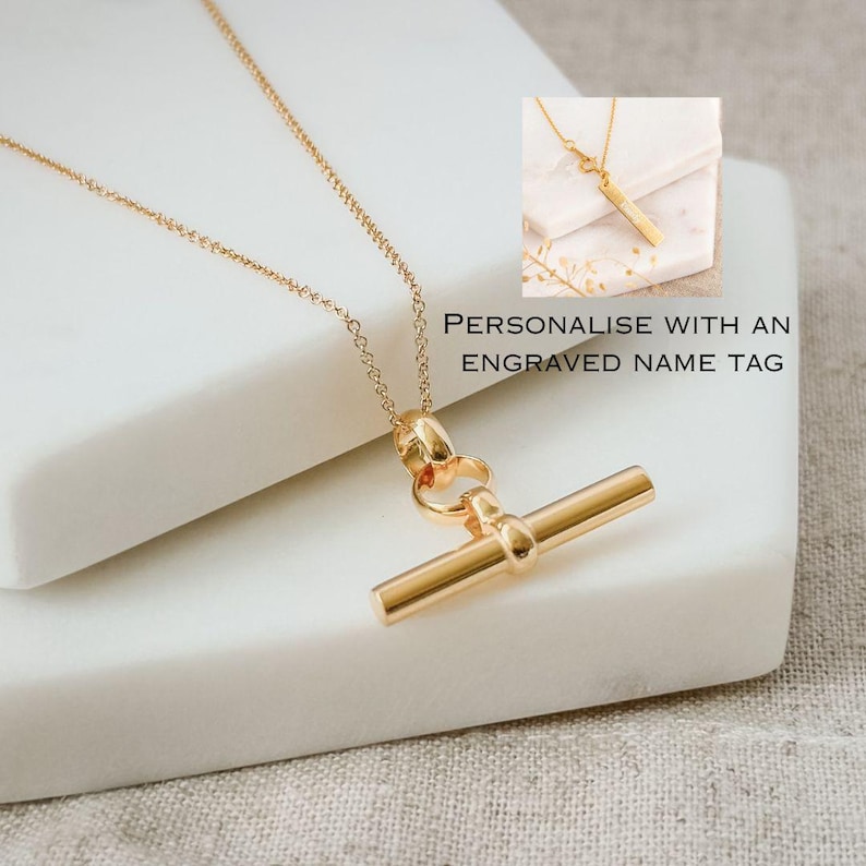 May include: Gold necklace with a toggle bar pendant. The pendant is a simple, straight bar with a small round accent at the top.  "Personalise with an engraved name tag" is written below the necklace.
