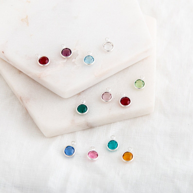 May include: Assortment of colorful crystal charms with silver settings. The charms are arranged on a white marble surface.
