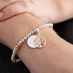 Könnte beinhalten: Ein silbernes und roségoldenes Perlenarmband mit einem Lebensbaum-Anhänger. Der Anhänger ist aus Roségold und hat ein filigranes Design.