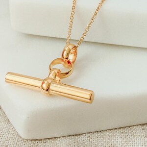 May include: A gold necklace with a toggle bar pendant. The pendant is a simple, straight bar with rounded ends. The chain is a delicate, linked chain.