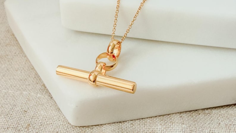 May include: A gold necklace with a toggle bar pendant. The pendant is a simple, straight bar with rounded ends. The chain is a delicate, linked chain.