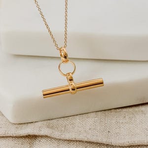 May include: A gold necklace with a simple bar pendant. The bar is attached to a small ring and a chain.