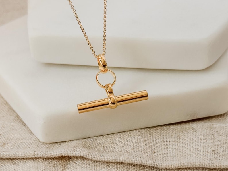May include: A gold necklace with a simple bar pendant. The bar is attached to a small ring and a chain.