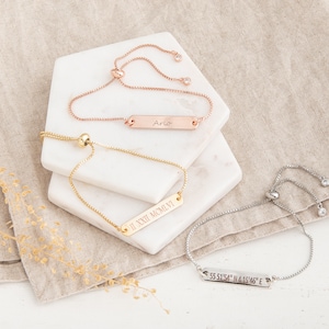 May include: A selection of personalised bar bracelets in rose gold, gold, and silver. Each bracelet has a rectangular bar with engraved text, including a name, Roman numerals, and coordinates. The bracelets are displayed on white marble and a neutral linen cloth.