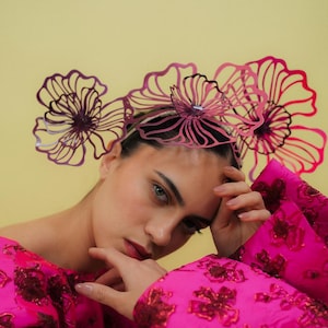 May include: A woman wearing a magenta floral headpiece and a matching long-sleeved dress. The headpiece features three large, stylized flower shapes. The dress has a textured floral pattern and voluminous sleeves. The background is a solid yellow.
