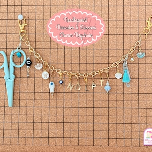 May include: Gold chain keychain with various charms, including a pair of blue scissors, a white seashell, a blue butterfly, a blue evil eye charm, and gold letter charms spelling "NDLPT". The keychain also features a blue and white dress charm and a white pearl.