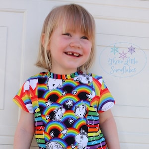 May include: A vibrant t-shirt with a rainbow and unicorn pattern. The design includes rainbows, stars, and white unicorns. The sleeves and neckline have rainbow stripes. The text "Three Little Snowflakes" is visible on the shirt.