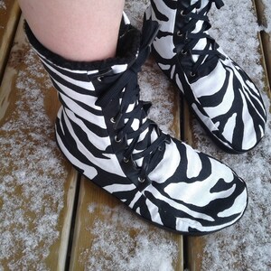 May include: A pair of black and white zebra print boots with black laces. The boots have a faux fur lining and are sitting on a wooden surface covered in snow. The boots are a high-top style.