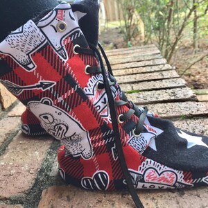 Może przedstawiać: Czerwono-czarne buty za kostkę z wzorem w kratę i białymi kreskówkowymi niedźwiadkami, sercami i gwiazdami. Buty mają czarne sznurowadła i białą strzałkę z czerwoną końcówką.