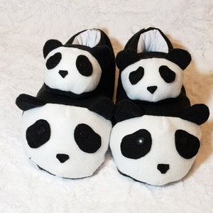 May include: A pair of panda bear slippers. The slippers are white with black accents, including the ears, eyes, and nose. The slippers are designed to resemble the face of a panda bear.