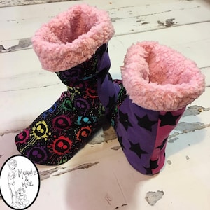 May include: A pair of colorful, fuzzy slippers with a pink fleece lining. One slipper has a black background with rainbow paint splatters and skull designs. The other slipper has a purple background with black stars.
