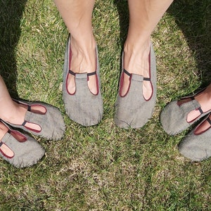 Puede incluir: Zapatillas de lino gris con diseño de tira en T y ribete burdeos. Las zapatillas se muestran sobre una superficie de hierba. Los zapatos tienen una puntera redondeada y un tacón pequeño. La tira en T es negra.