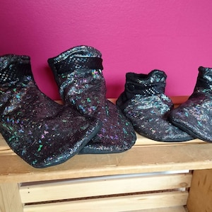 May include: Four pairs of black, sparkly boots are displayed on a wooden crate against a magenta background. The boots have a textured, iridescent finish with a black mesh detail at the top. The boots vary in size.