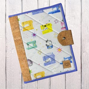 May include: Quilted notebook cover with a white background featuring colorful sewing machines and star patterns. The cover has a cork spine and a cork closure with a metal snap. The edges are trimmed with a blue binding.