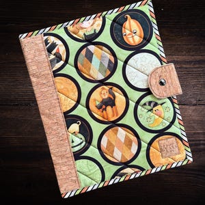 May include: A quilted journal cover with a light green background and circular Halloween-themed illustrations. The cover features a cork spine and a wooden button closure. The illustrations include pumpkins, cats, and moons. The cover is bordered with a striped pattern.