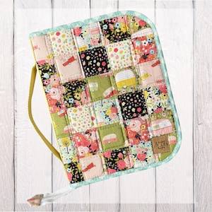May include: A rectangular quilted fabric case featuring a patchwork design of floral patterns and vintage camper illustrations in shades of pink, green, black, and yellow. It has a light blue trim, a yellow side handle, and a small brown label.