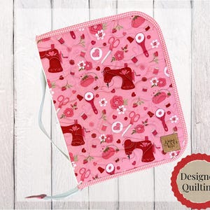 May include: A quilted pink fabric case featuring a repeating pattern of red sewing machines, scissors, rotary cutters, and pink roses. The case has a white zipper, a white carrying strap, and a small brown rectangular label with text.