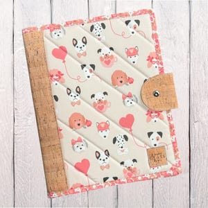 May include: Quilted notebook cover with a cork spine and closure. The cover features a cream background with a repeating pattern of cartoon dogs, hearts, and bows in pink and black. The edges are trimmed with a pink floral pattern.