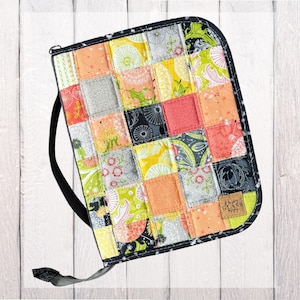 May include: A rectangular zippered case featuring a patchwork design of various fabric squares in shades of yellow, orange, green, gray, and black. The case has a black fabric handle and a small brown square label with the text A Sewing Glass.