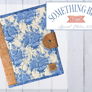 May include: A quilted fabric notebook cover featuring a blue and cream floral toile pattern, accented with a brown cork spine and a matching cork tab closure with a metal snap. The cover includes a small square label and text reading SOMETHING BLUE Tilda Special Edition 2026.