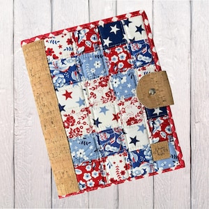 May include: A quilted fabric notebook cover featuring a patchwork pattern of red, white, and blue floral and star prints. The cover includes a vertical cork spine, a matching cork tab closure with a metal snap, and a small square cork label.