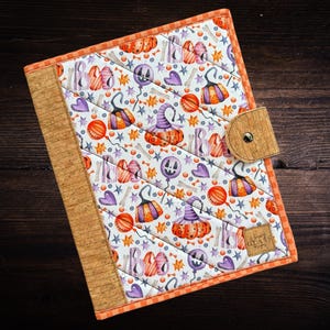 May include: A quilted Halloween-themed journal with a cork spine and closure. The cover features a repeating pattern of pumpkins, witch hats, bones, and balloons in orange, purple, and white. The journal is bordered with orange fabric.