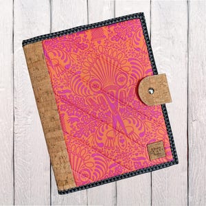 May include: A quilted journal cover with a vibrant orange and pink floral pattern. The cover features a cork spine and a cork closure with a silver snap. The edges are trimmed with a black and gray patterned fabric. A small tag is attached to the cover.