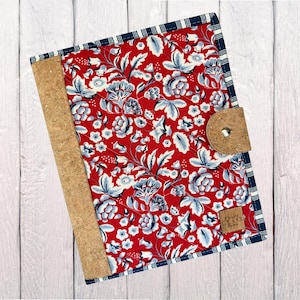 May include: A fabric-covered journal with a red floral pattern and a cork spine. The journal has a cork closure and a navy blue and white striped border. The journal is on a white wooden surface.