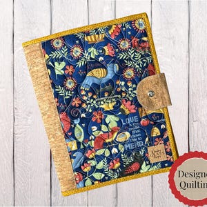 May include: A rectangular fabric cover featuring a dark blue background with a colorful floral and bird pattern, quilted with a gold border. It includes a cork spine and a cork tab closure with a metal snap. The text reads Love is the needle that knows how to mend.