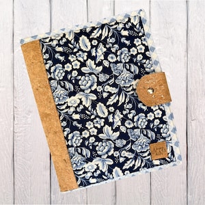May include: A floral patterned journal cover with a cork spine and closure. The cover features a navy blue background with white and light blue floral designs. A small cork tab secures the journal closed.