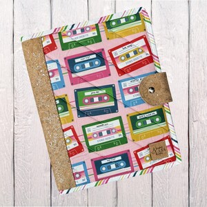 May include: A quilted notebook cover featuring a pink background with colorful cassette tape designs. The cover has a cork spine and a cork closure with a button. The edges are trimmed with a striped pattern. A small tag with text is visible.