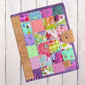 Może przedstawiać: Kolorowa patchworkowa okładka na dziennik z korkowym grzbietem i korkowym zapięciem. Okładka posiada kwadraty materiału w różnych wzorach i kolorach, w tym różowe, fioletowe, zielone i kwiatowe wzory. Etykieta głosi "Whimsical Glass Designs."